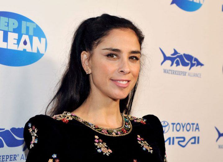 A woman standing in front of a white wall with blue Waterkeeper Alliance logos on it. She is Sarah Silverman.