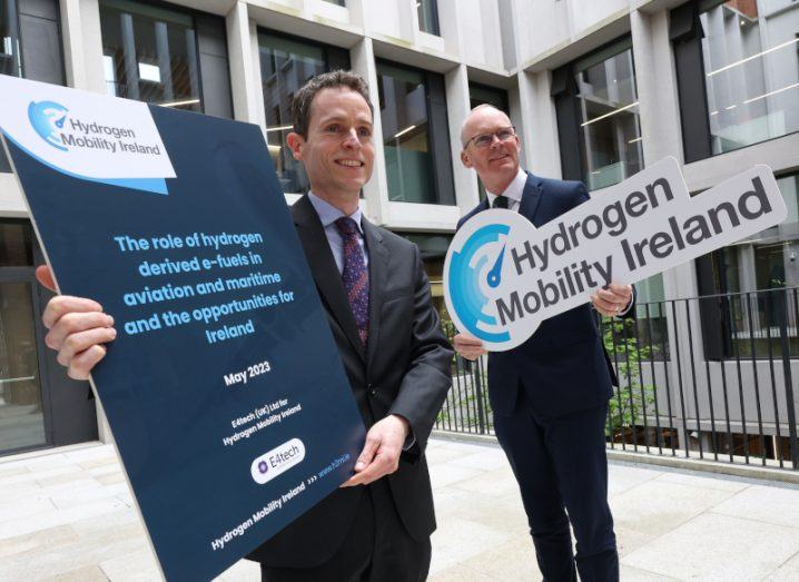 Two men holding signs for the Hydrogen Mobility Ireland report. They are standing outside a building.