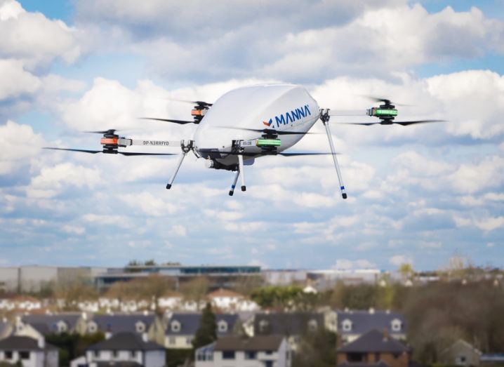 A white drone pictured against a mostly sky background, flying over suburban houses.