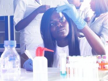 A young woman looking at a test tube in a lab environment with other biopharma workers standing behind her.