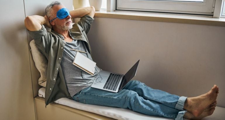 Man wearing an eye mask resting while sitting in a window seat in a relaxed pose with a laptop and notebook on his lap.