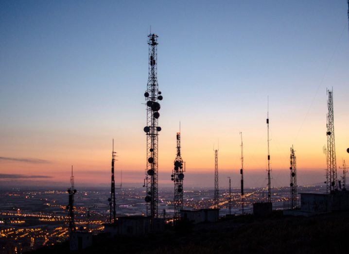 Multiple communication towers in front of an evening sky, with a sunset in the background and building lights below.
