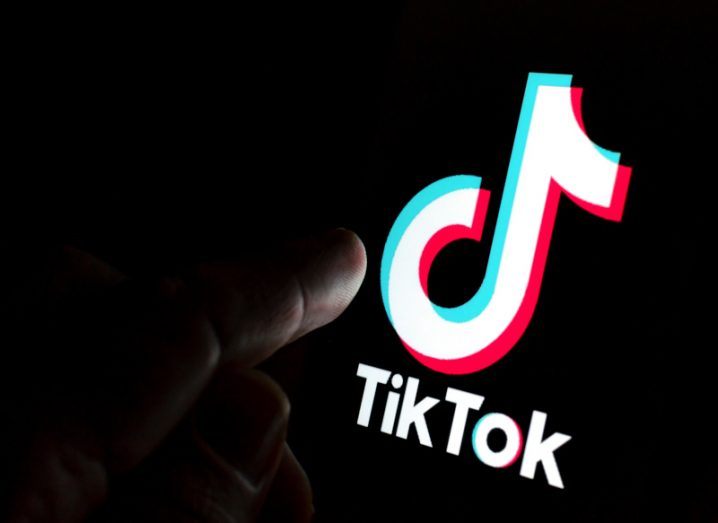The TikTok logo on a screen in a dark room, being touched by a person's finger.