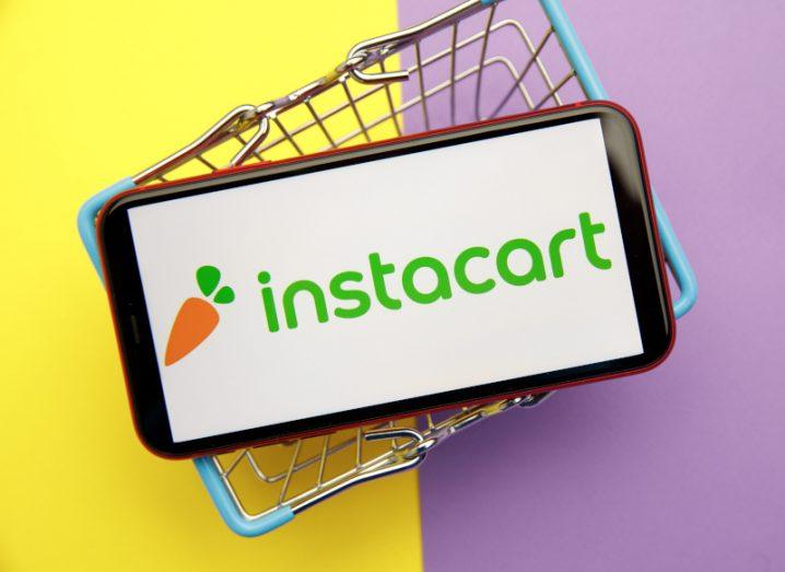 The Instacart logo on a smartphone screen. The phone is on a small shopping cart, on a yellow and purple surface.