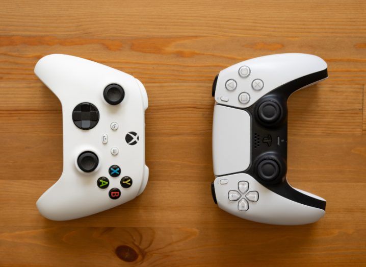 Two console controllers facing opposite each other on a wooden table. The controllers are for the Xbox and the Playstation, two consoles made by Microsoft and Sony.