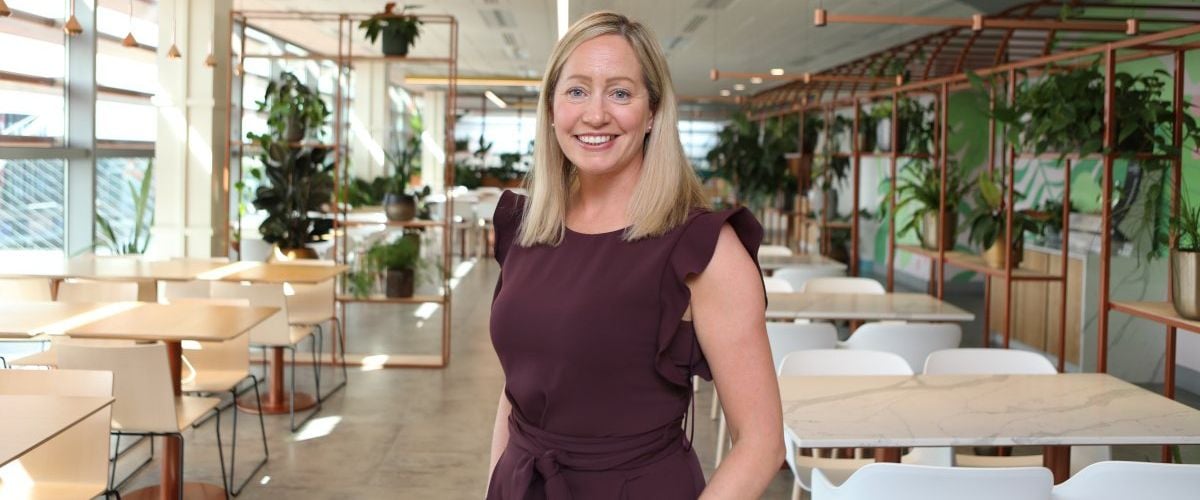 A woman wearing a maroon dress stands smiling in a brightly lit office setting with various tables and plants in the background. She is Aisling Curtis, market leader at PwC's strategic alliances team.