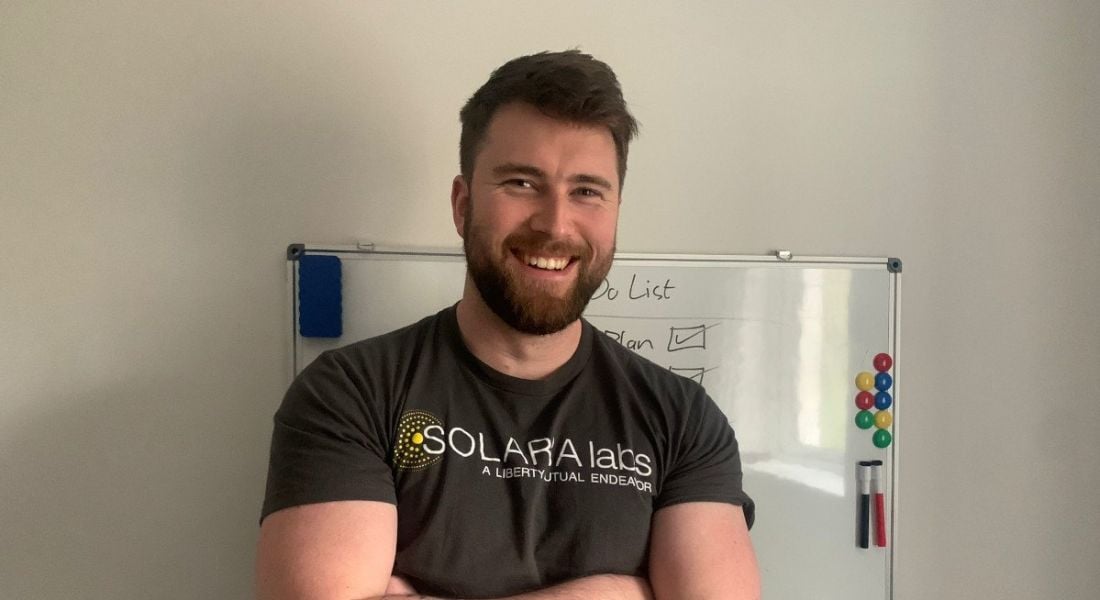 A man stands smiling with his arms folded in front of a whiteboard stuck on a wall. He is Alan Quigley, a principal software engineer at Liberty IT.