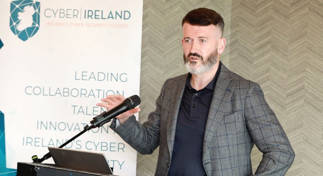 A man standing at a podium speaking about cybersecurity. There is a board behind him with Cyber Ireland branding.