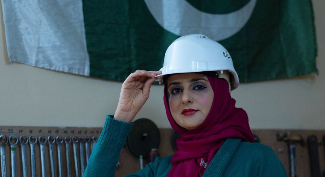 A close-up of Dr Sarah Qureshi in a white hard hat with her hand on the rim of the hat as she looks at the camera.