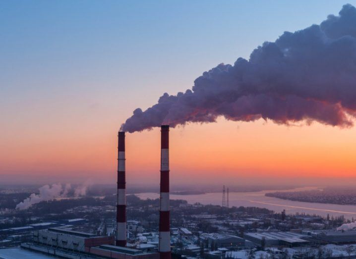 An industrial area with two large red and white chimney stacks releasing harmful emissions into the atmosphere at sunset.