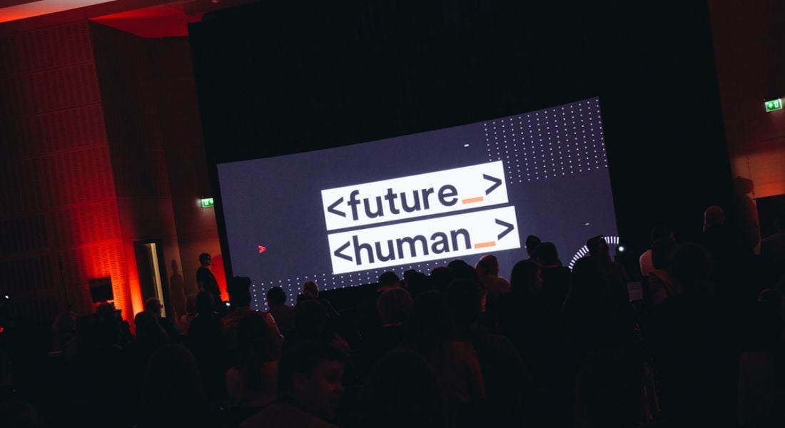 A darkened room with a large seated crowd in front of a large screen with the Future Human logo on display.