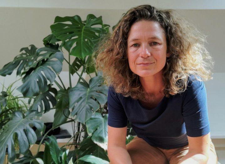 Marije Schaafsma, a woman with curly hair sits leaning forward with her elbows rested on her thighs. She is wearing a dark blue t-shirt and there is a large green plant behind her.