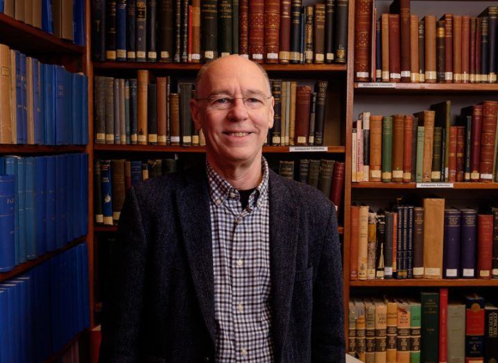 Prof Simon Jeffery wears a checked shirt and a woollen blazer and smiles at the camera. He is standing in front of shelves with very old books on them.
