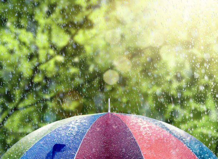 A colourful umbrella at the bottom of the picture with green leaves in the background and rain with sun shining through leaves at the right.