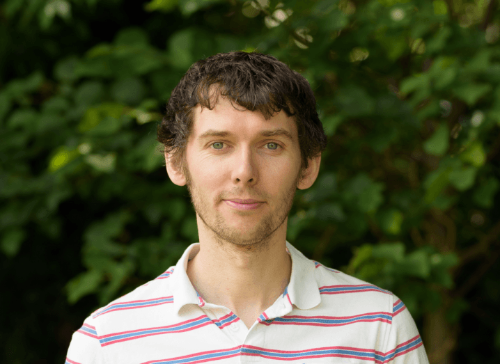 Eoin Hinchy, CEO of Tines, standing in front of foliage. He has slightly curly, dark hair and is wearing a white polo shirt with red and blue stripes.