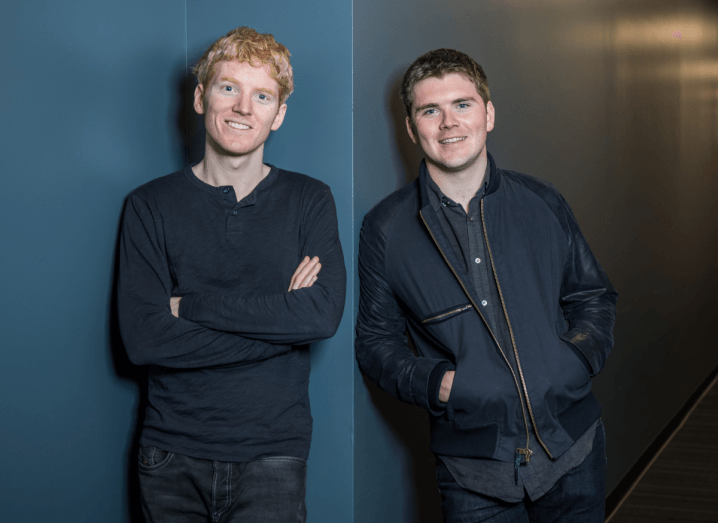 Stripe's founders, the Collison brothers, standing in front of a dark blue wall. One has red hair and a navy long-sleeved shirt on, with his arms crossed. The other has brown hair and a navy jacket, with his hands in his pockets.