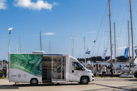 The TREC EMBL van parked at a dock with yachts behind it and a blue sky and small white clouds in the background. The side door of the van is open with shelves visible inside.